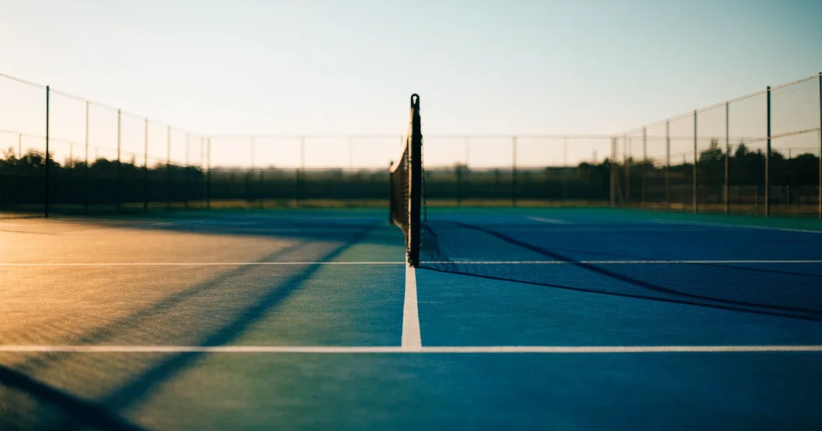 Ein leerer Tennisplatz mit Hartplatzbelag in der Morgendämmerung, das Netz im Gegenlicht und lange Schatten der Linien.