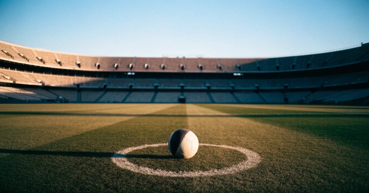 Ein Fußball liegt auf dem Anstoßpunkt eines leeren Rasenplatzes, im Hintergrund eine große Stadiontribüne im Abendlicht.