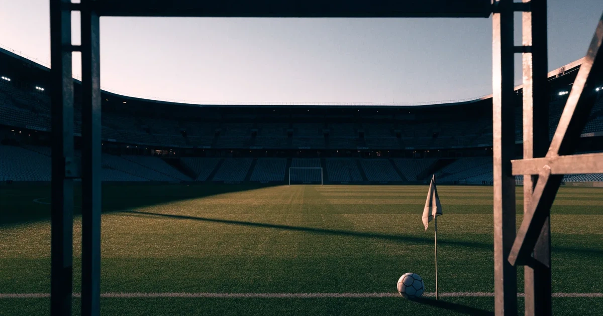 Blick durch ein Stadiontor auf einen grünen Rasen im Abendlicht, ein Ball ruht an der Eckfahne, die Tribünen sind leer.