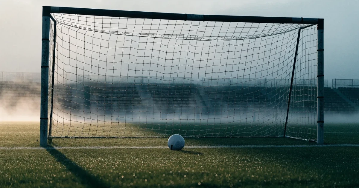 Ein Fußballtor mit gespanntem Netz steht auf einem leeren Rasenplatz, davor ein einzelner Ball, Morgendunst liegt über dem Feld.