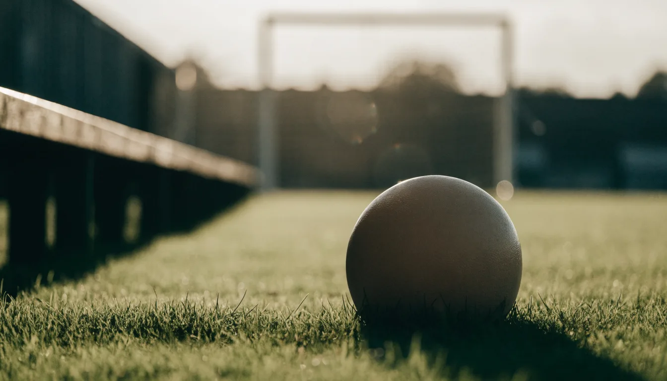 Ein Fußball liegt im Gras am Spielfeldrand, dahinter eine leere Bank und ein Tor im Gegenlicht.