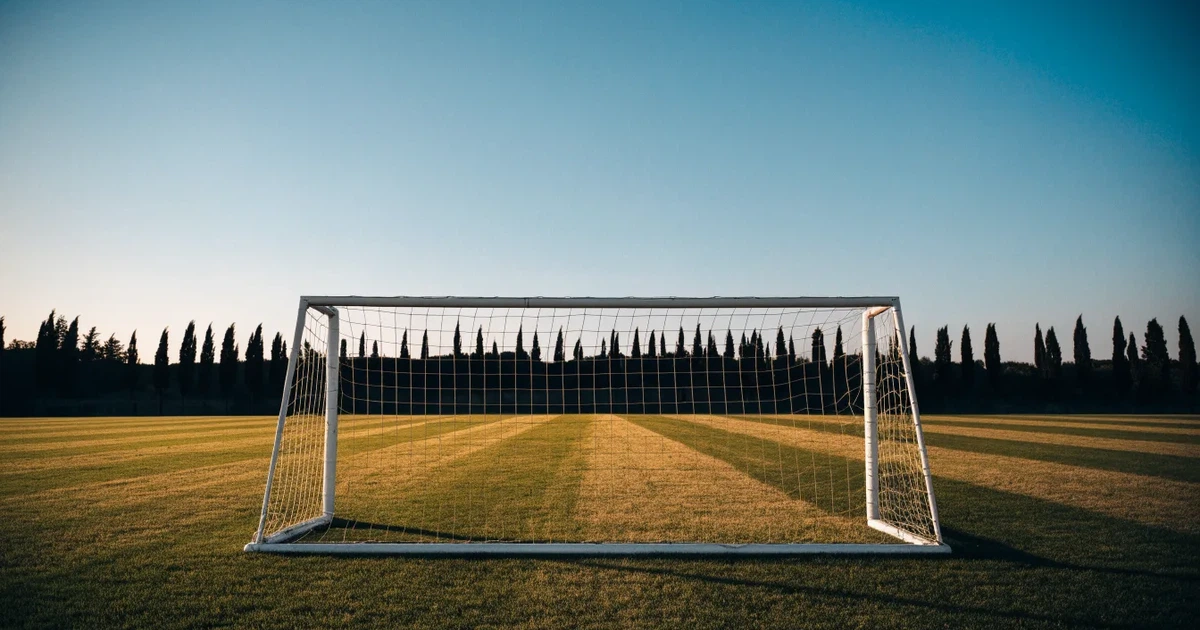 Ein Fußballtor mit Netz auf einem leeren Platz im Abendlicht, der Rasen frisch gemäht, im Hintergrund Zypressen.