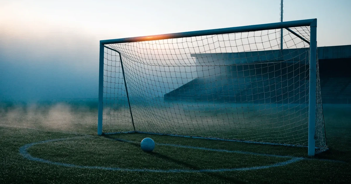 Ein Fußballtor mit Netz steht auf einem leeren Platz im Nebel der Morgendämmerung, der Rasen ist taufeucht.