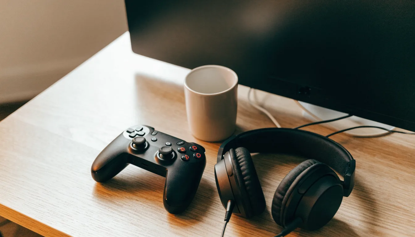 Ein Gamepad und ein Headset liegen auf einem Schreibtisch neben einer Tasse, der Bildschirm im Hintergrund ist ausgeschaltet.