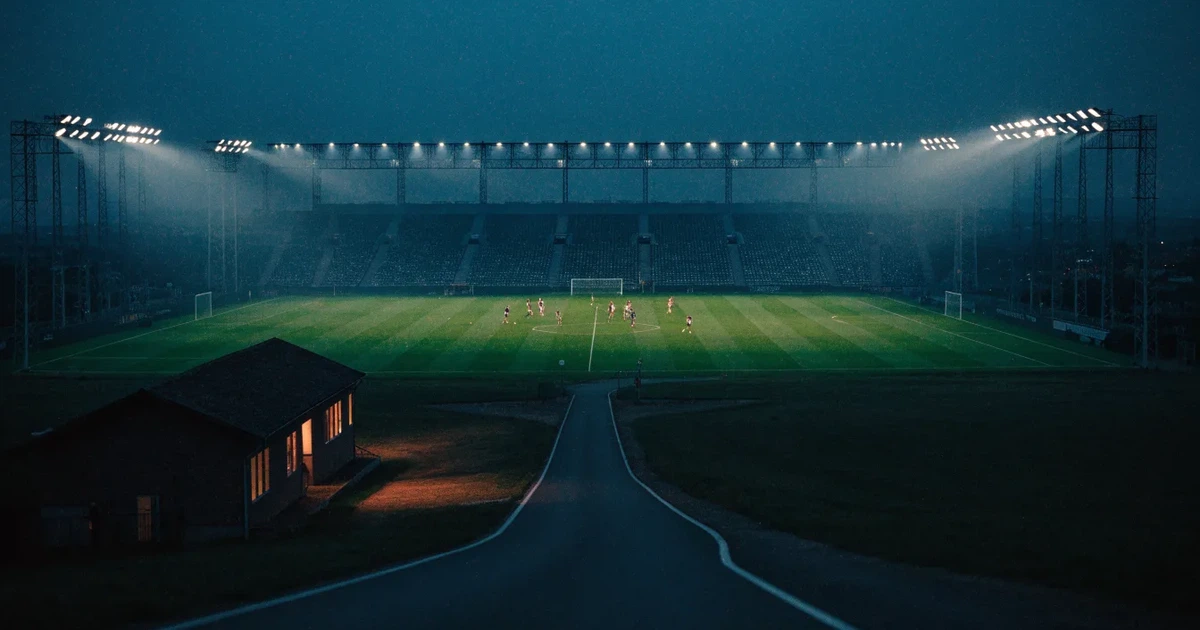 Ein Fußballstadion bei Flutlicht aus der Ferne gesehen, davor eine Straße und leuchtende Fenster eines Nebengebäudes.