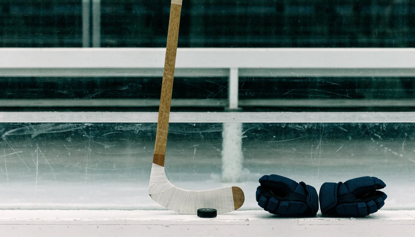 Ein Eishockeyschläger lehnt an einer Bande, daneben liegen ein Puck und ein Paar Handschuhe in einer Eishalle.