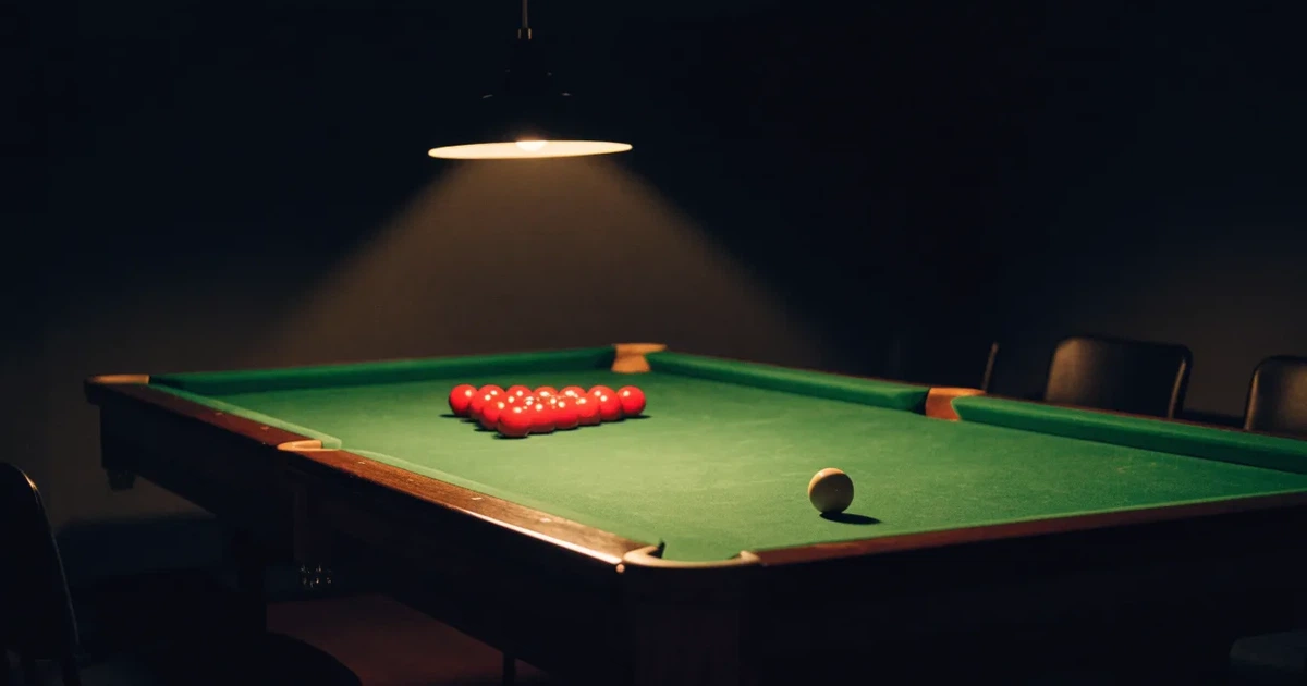 Ein Snookertisch mit grünem Tuch und aufgebauten roten Kugeln in einem abgedunkelten Raum unter einer hängenden Lampe.