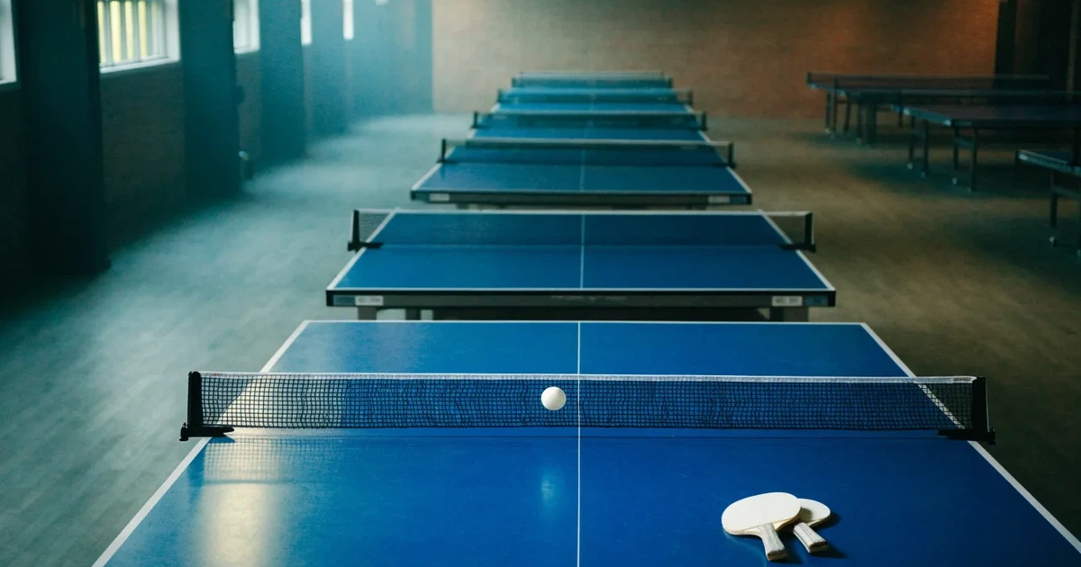 Eine Tischtennishalle aus erhöhter Perspektive mit mehreren blauen Tischen in Reihen unter gedämpftem Hallenlicht, ohne Personen.