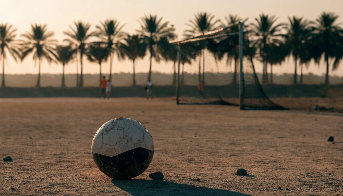 Ein Fußball liegt auf einem staubigen Bolzplatz mit Palmen im Hintergrund in warmem Abendlicht.