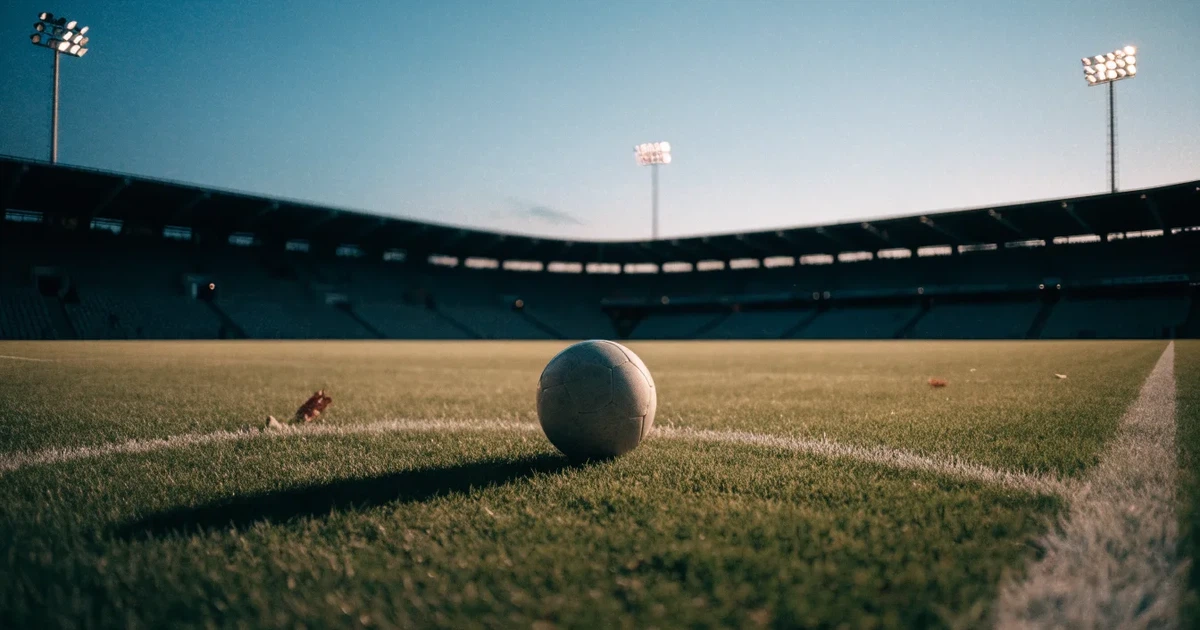 Ein Fußball liegt auf dem Anstoßpunkt eines leeren Rasenplatzes, im Hintergrund eine Tribüne und Flutlichtmasten in der Abenddämmerung.