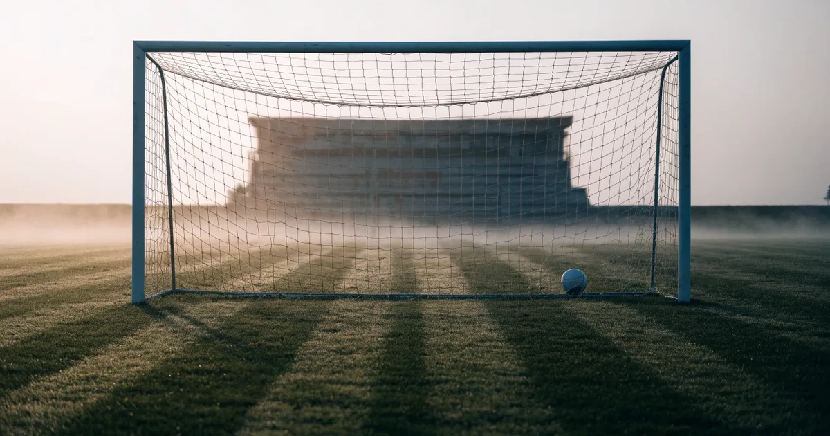 Ein Fußballtor mit Netz steht im Morgennebel auf einem leeren Rasenplatz, im Hintergrund die Silhouette einer kleinen Tribüne.