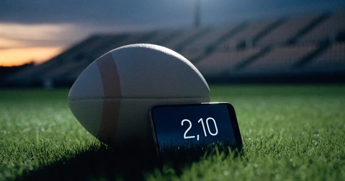 Ein American-Football-Ball liegt auf einem Rasenplatz unter Flutlicht, im Hintergrund eine große Tribüne in der Dämmerung.