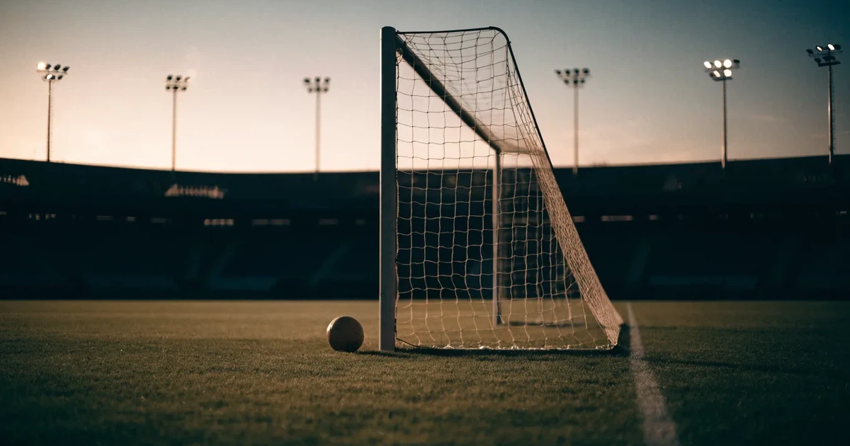 Ein Fußballtor mit Netz im Gegenlicht auf einem leeren Platz am Abend, davor ein Ball auf dem Rasen.