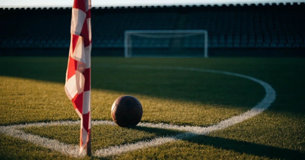 Ein Fußball liegt am Viertelkreis an der Eckfahne eines Rasenplatzes, im Hintergrund leere Tribünen in warmem Abendlicht.