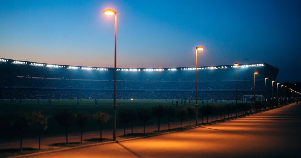 Ein großes Fußballstadion mit hell erleuchtetem Rund, aus der Ferne am Abend gesehen, davor eine breite Allee mit Laternen.