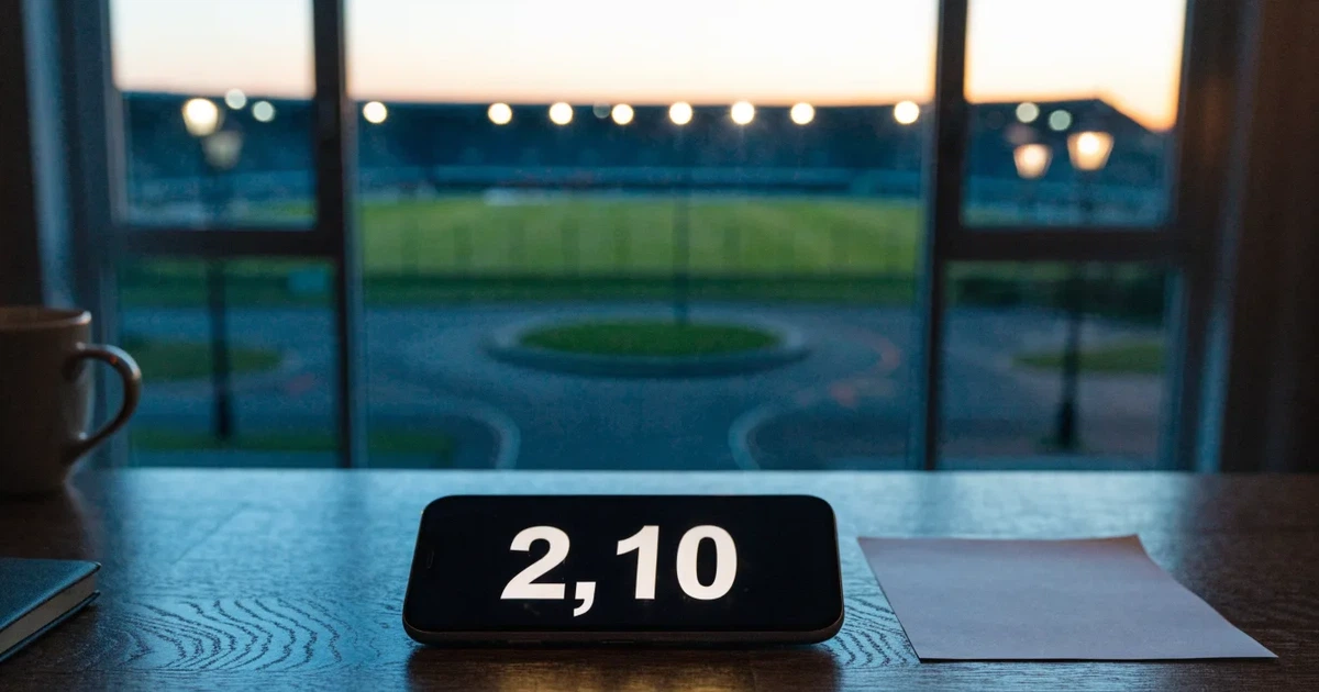 Ein Fußballstadion in der Dämmerung aus der Ferne, die Flutlichter gerade eingeschaltet, davor ein Kreisverkehr mit Laternen.